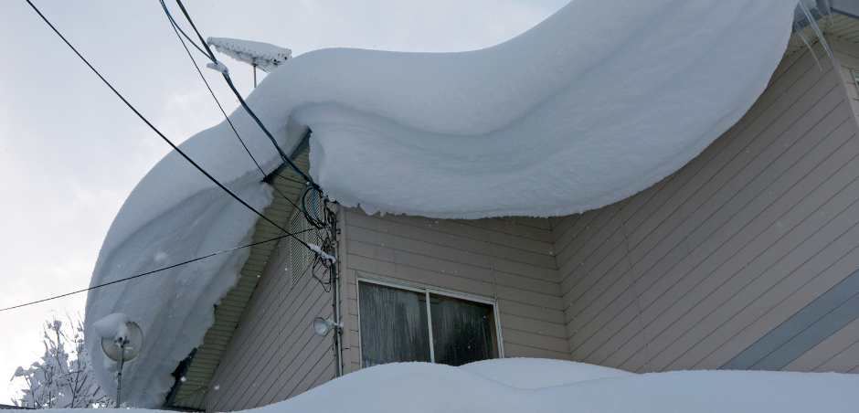 Massive snowdrift piled on a house roof, hanging over the eaves near a window.