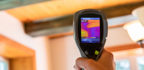 Handheld thermal camera scanning a ceiling with warm orange heat patterns displayed on the screen