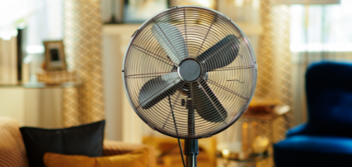 Black standing fan in a warmly lit living room with curtains and chairs in the background