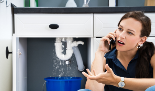 Woman on phone under a leaking sink cabinet, with water dripping into a blue bucket.