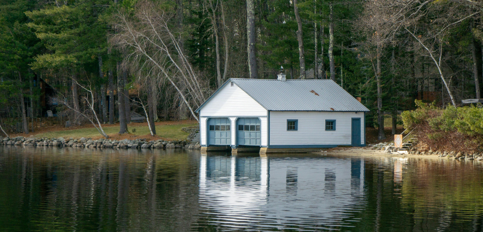 A white boathouse with two garage-style doors sits at the edge of a calm lake surrounded by a dense, wooded forest.