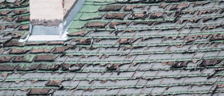 A severely damaged green asphalt shingle roof with missing material and exposed underlayment near a chimney.