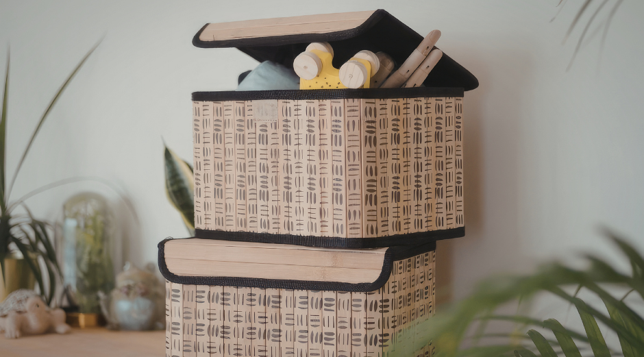 Stacked woven storage boxes with black hats and flowers on top, in a light room with plants in the background