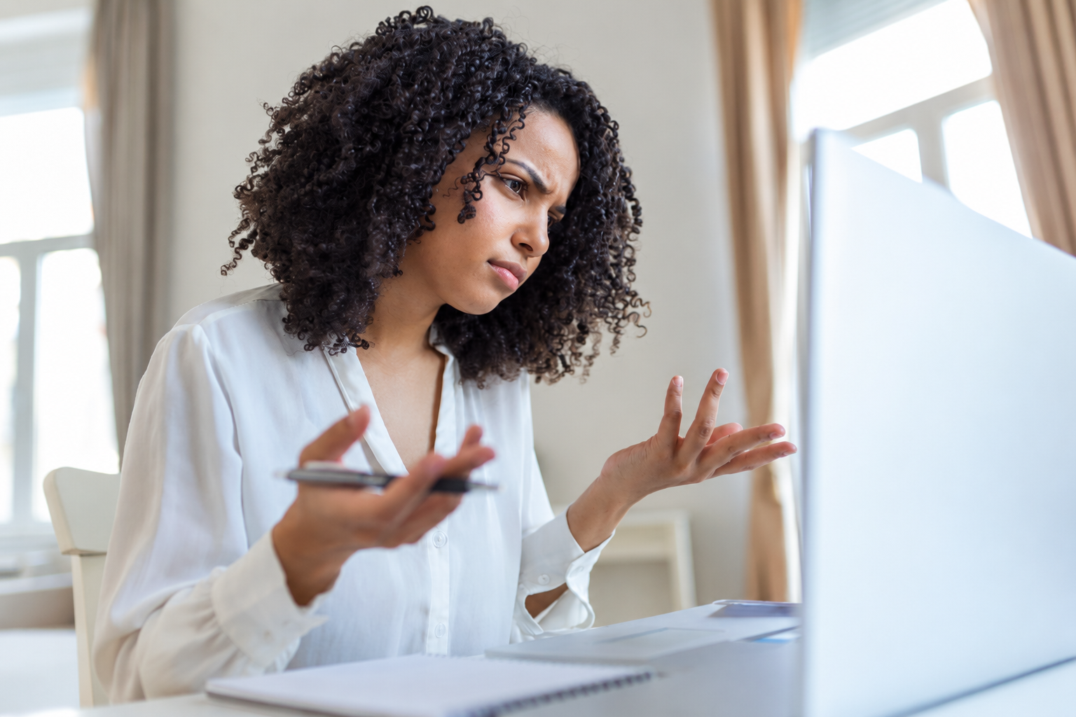 Woman in a white shirt speaking on a video call, gesturing with a pen at a laptop.