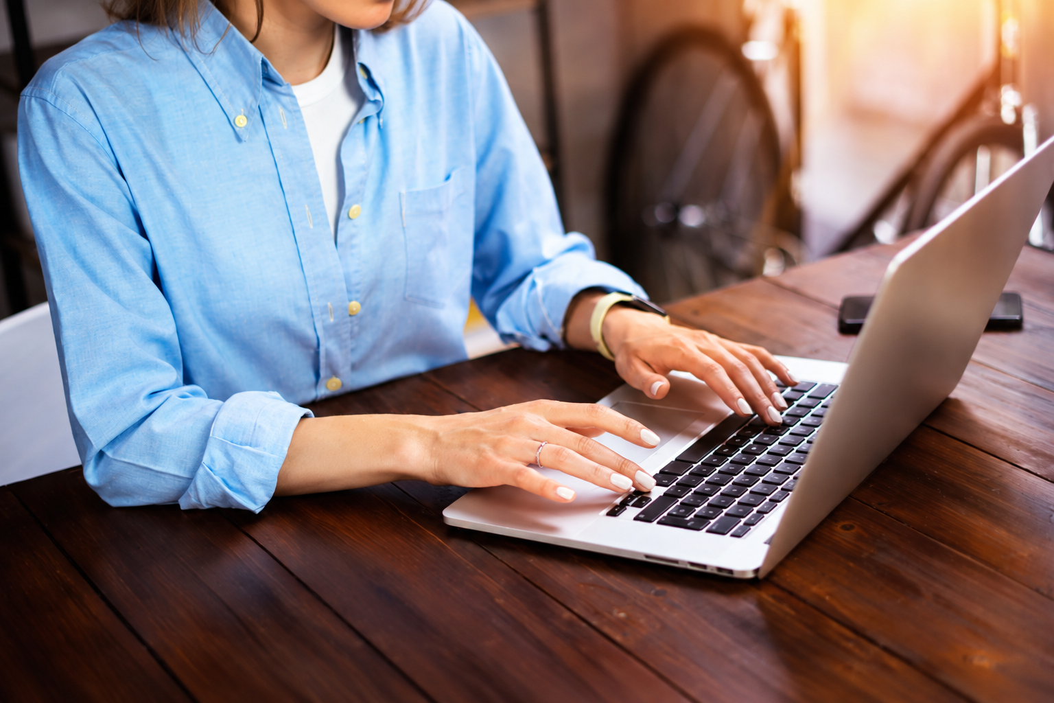 Person typing on a laptop at a wooden desk, wearing a light blue shirt.