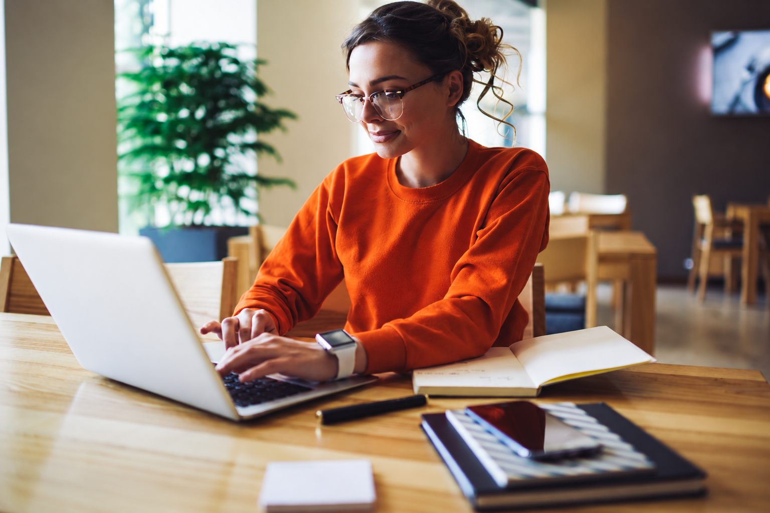 Person in an orange sweater working on a laptop at a desk in a bright room