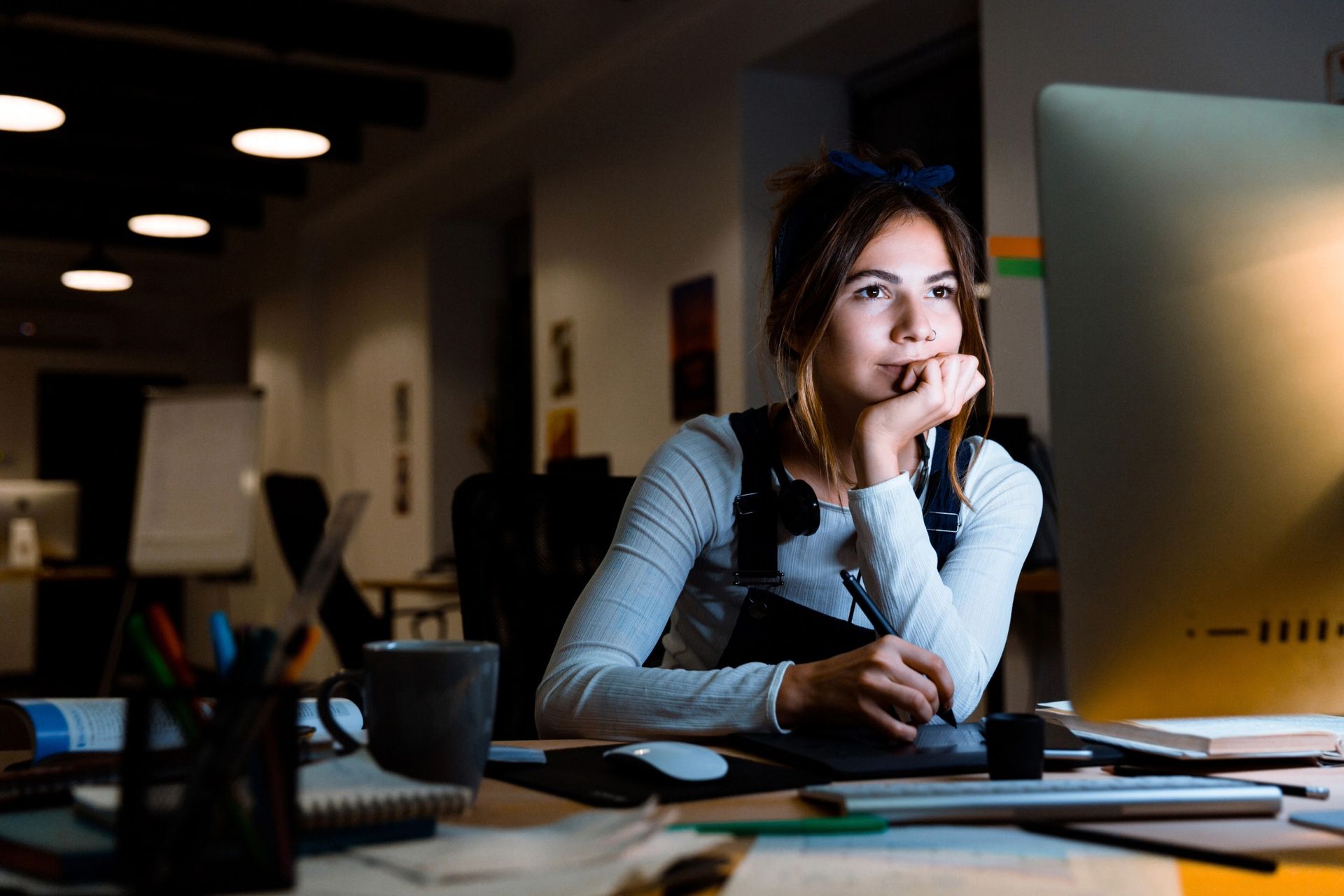 Woman sitting at a desk, focused on a computer screen, in a dimly lit office.