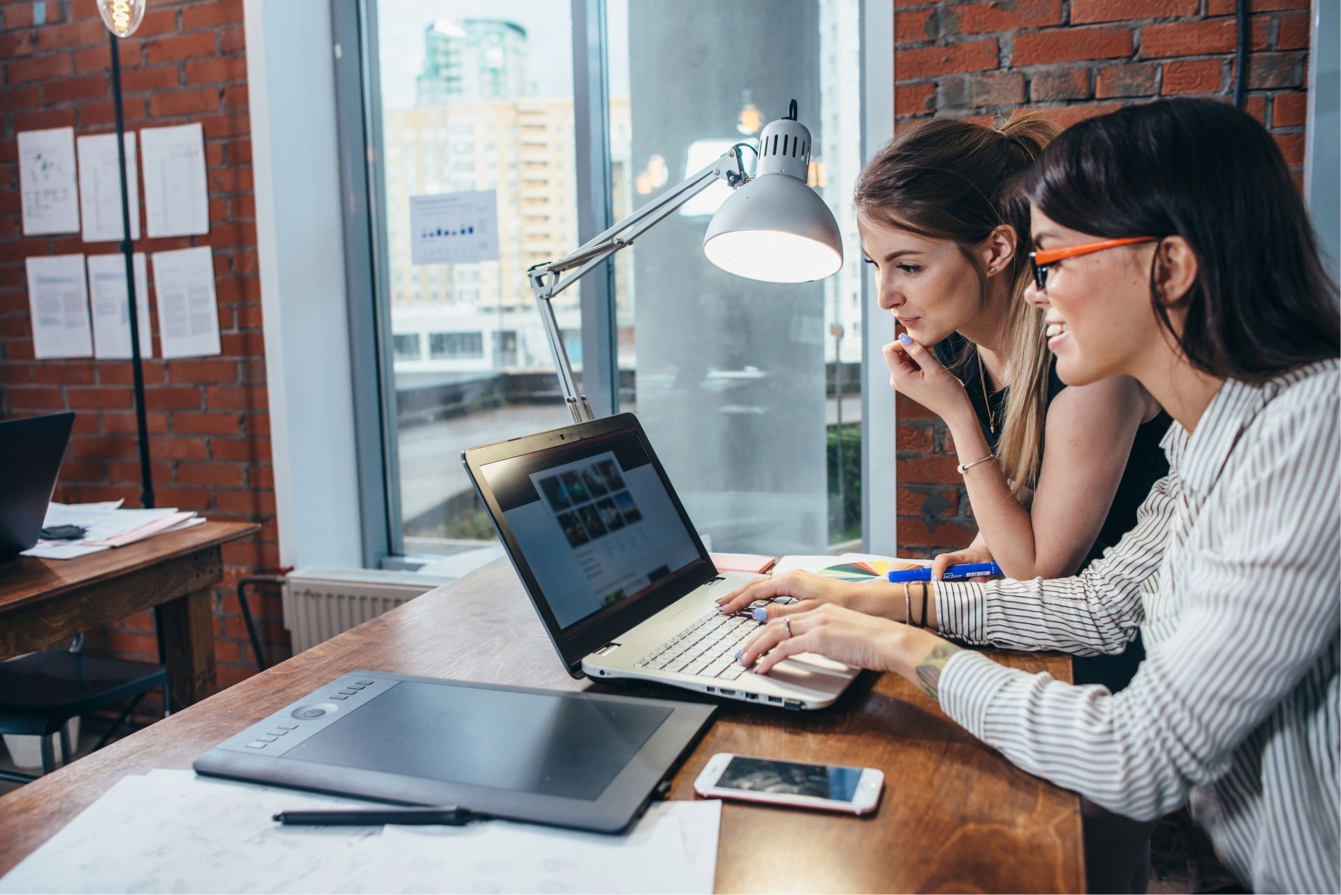 Two women looking at a laptop, one typing. Desk with tablet and phone, brick wall backdrop.