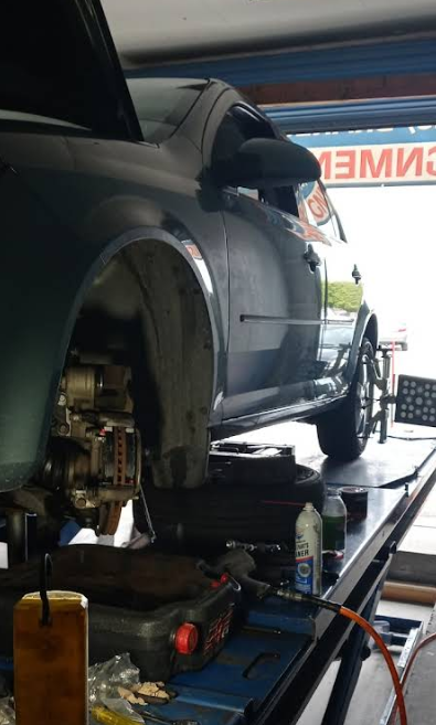 A dark car lifted on a garage rack for maintenance, with a brake rotor visible and alignment equipment on the rear wheel.