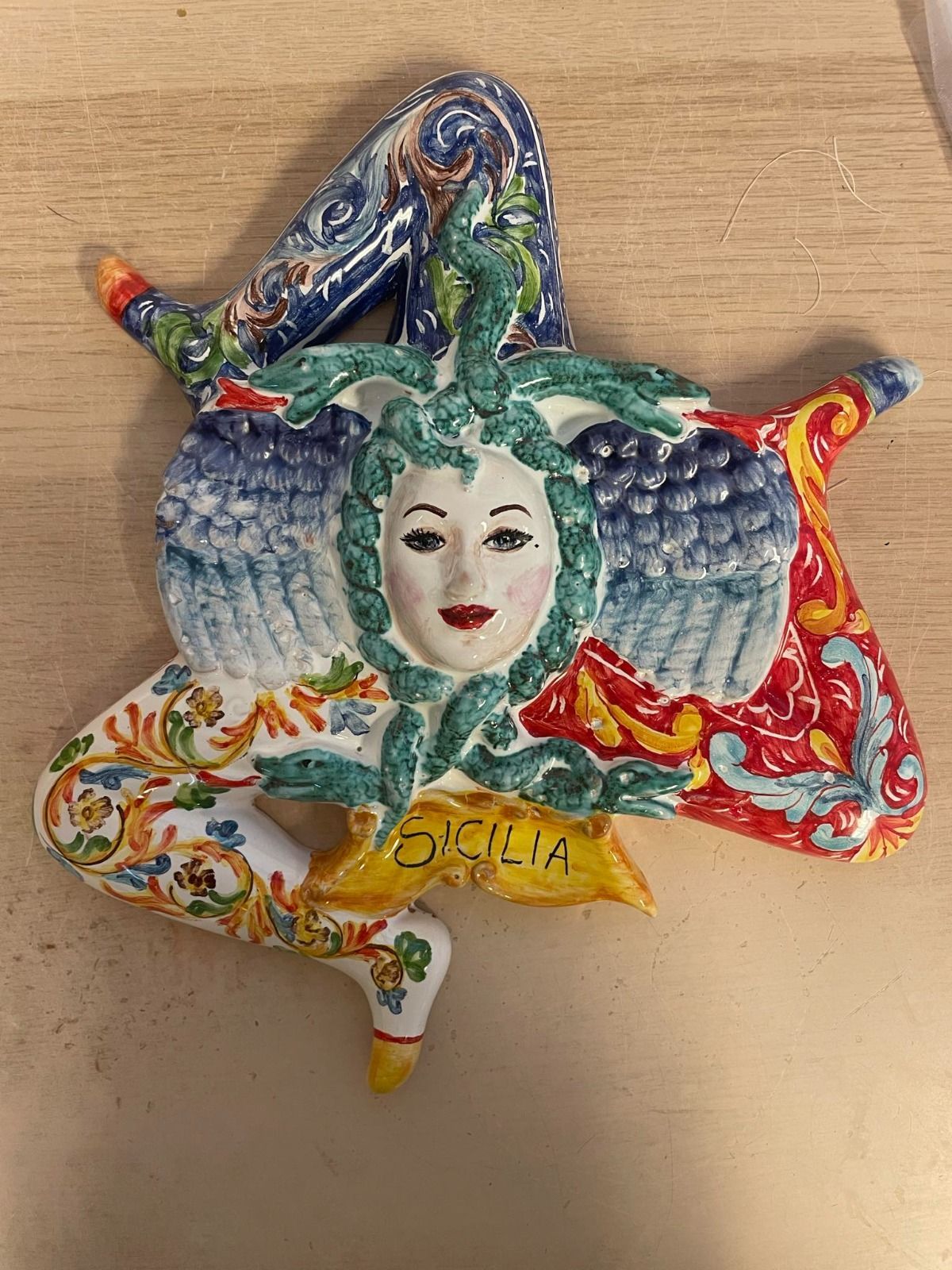 Emblema della Trinacria in ceramica, testa di donna con serpenti, tre gambe, motivi floreali colorati e scritta