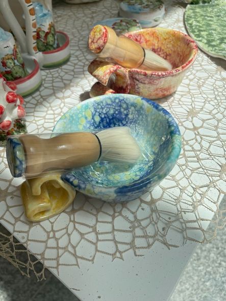 Ciotola da barba con pennello. La ciotola è blu, gialla e bianca, e sullo sfondo ci sono altri oggetti in ceramica.