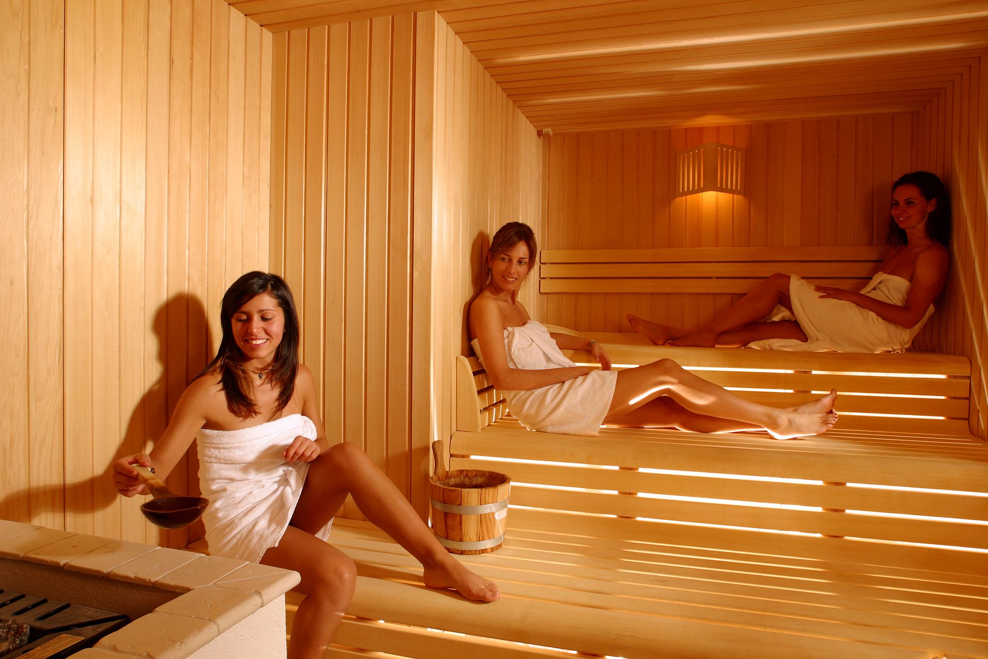 Drei in Handtücher gehüllte Frauen sitzen in einer Holzsauna.