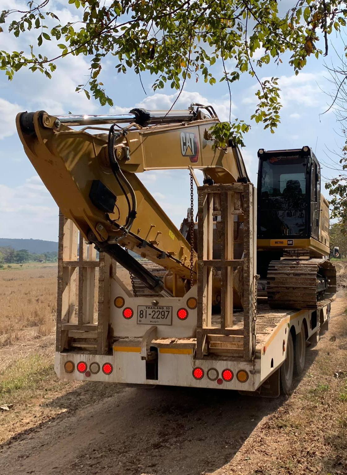 Yellow excavator on a flatbed trailer on a dirt road, viewed from behind under trees.