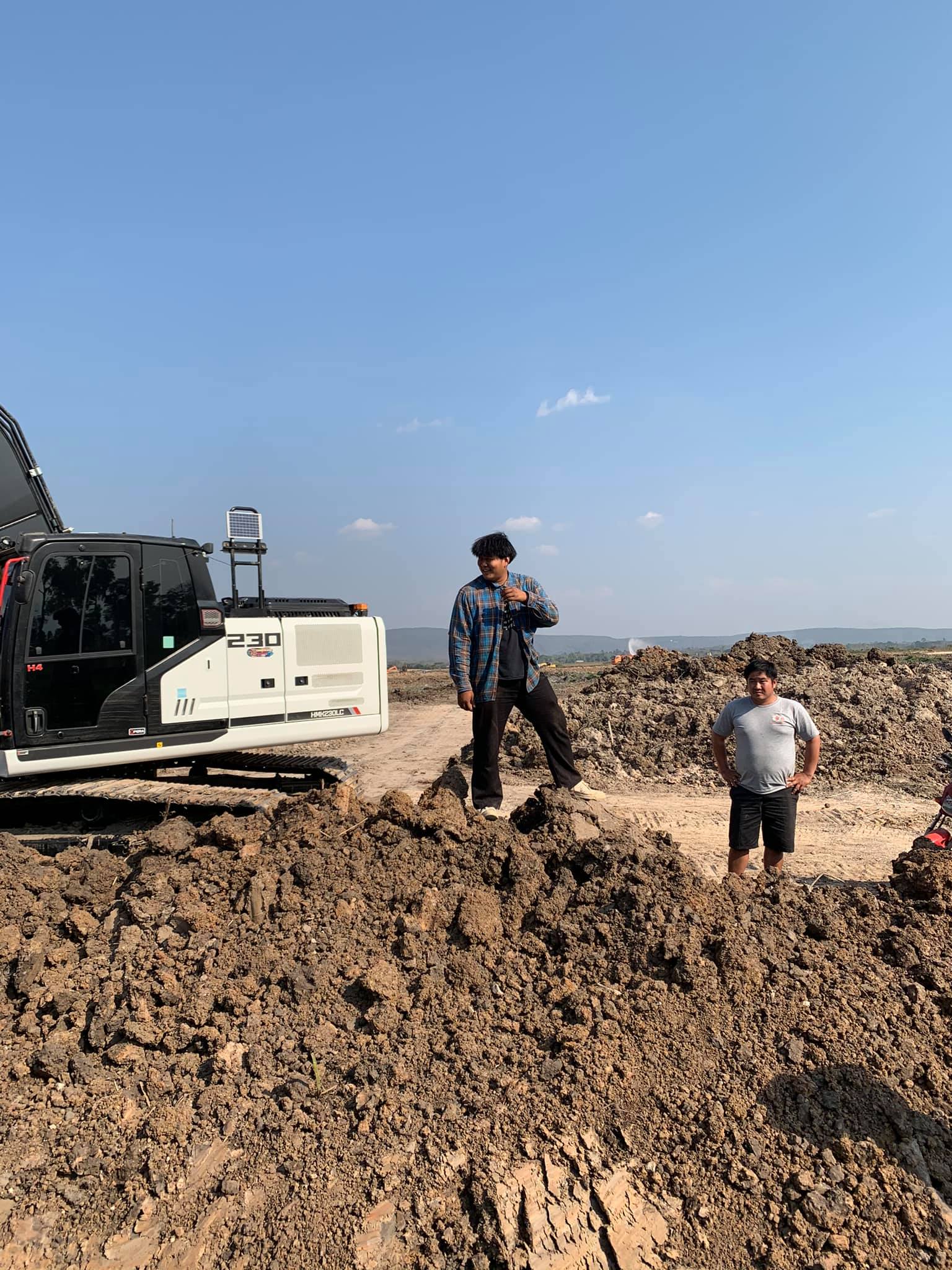 Two people standing on a large dirt mound beside a tracked machine under a clear blue sky