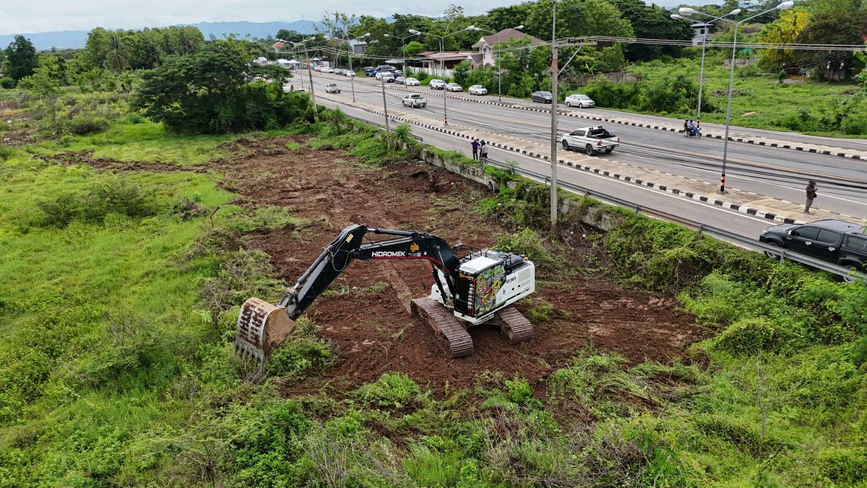 Excavator clearing a grassy roadside lot beside a highway under cloudy skies