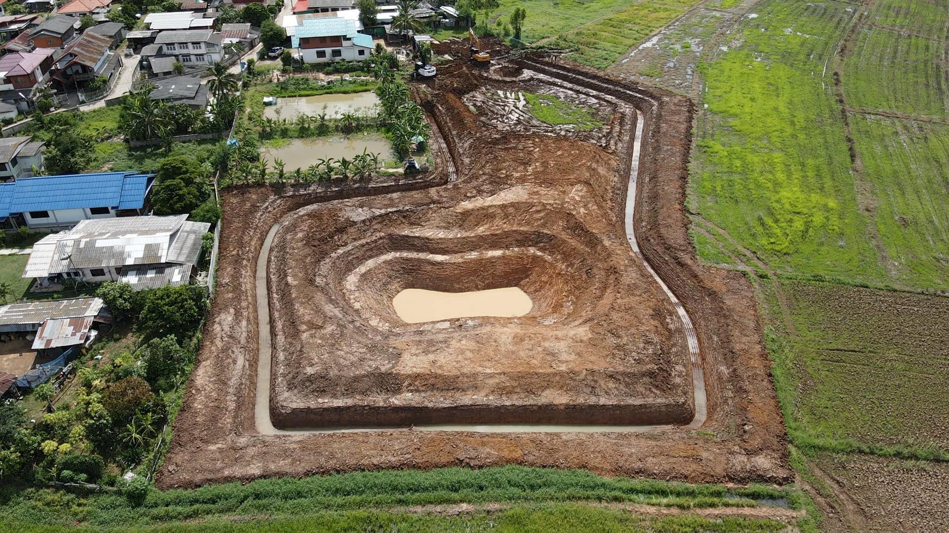 Aerial view of a terraced dirt excavation site with a central pond, bordered by farmland and houses.