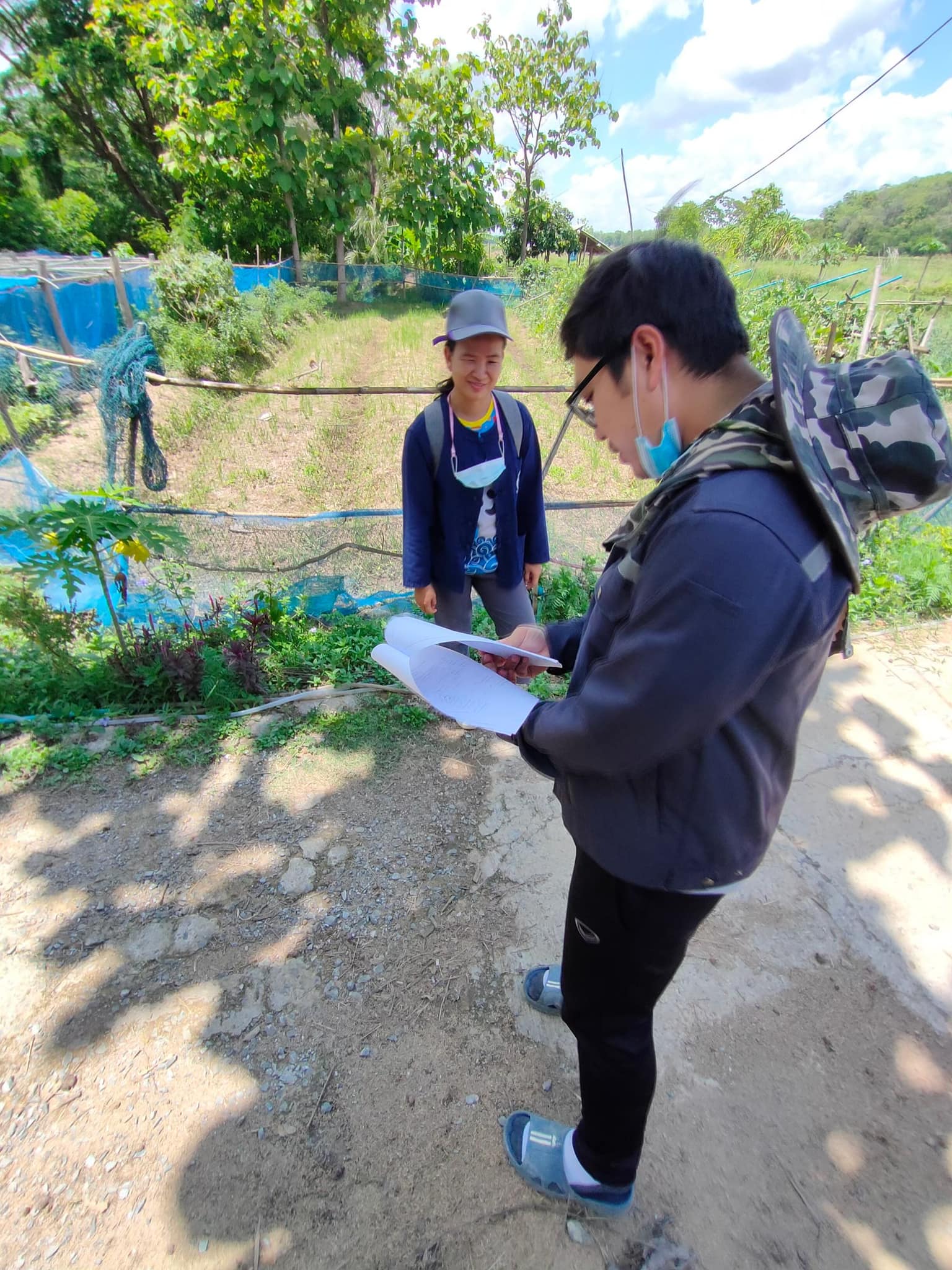 Two people outdoors by a garden path, one writing on a clipboard while the other stands nearby.