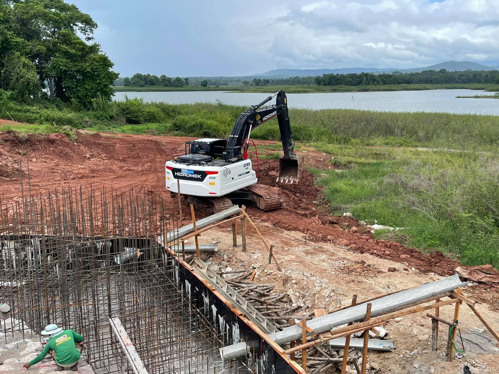 Excavator digging near a concrete foundation by a lake, with rebar and workers at a construction site.