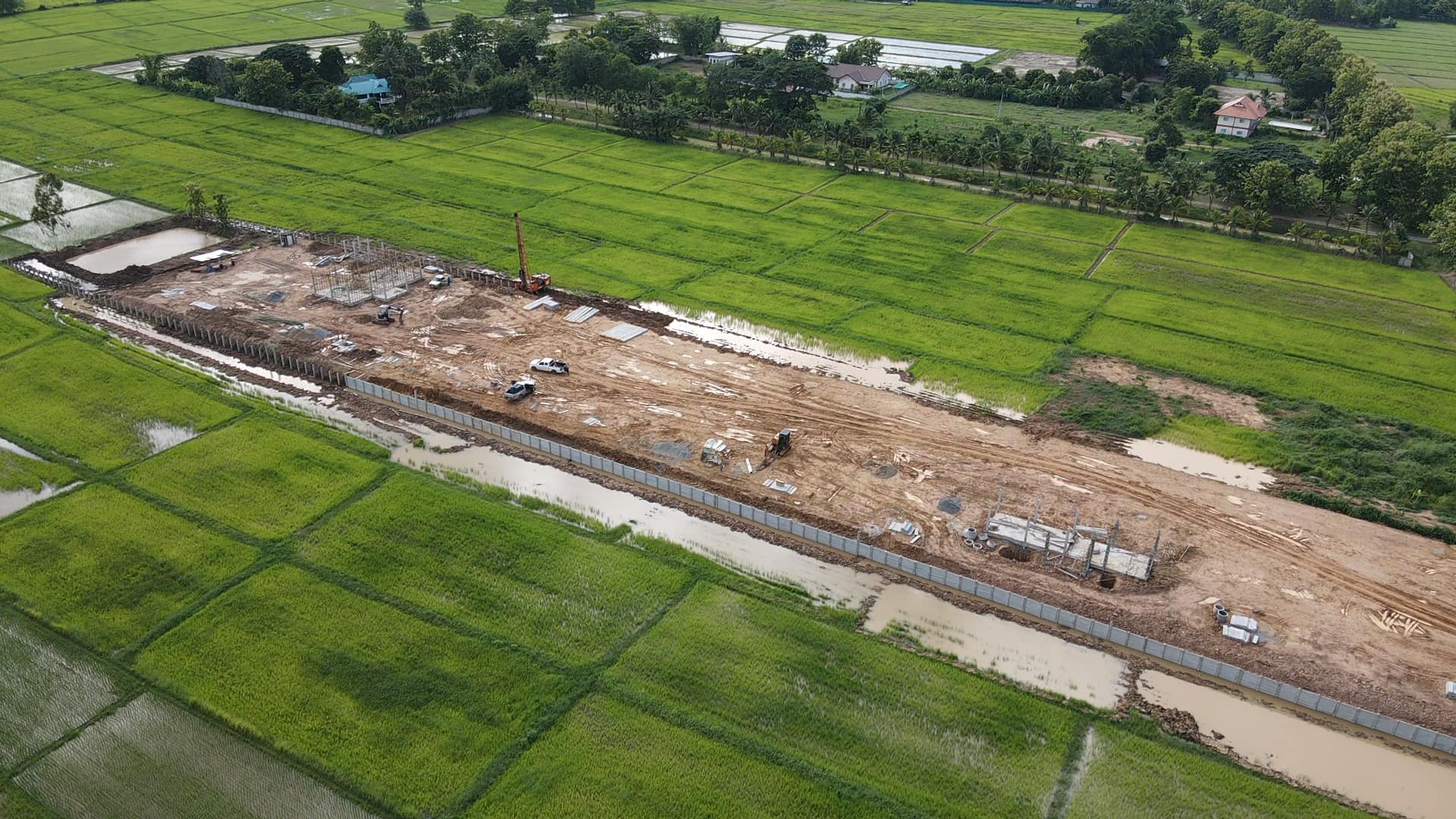 Aerial view of a rural construction site cutting through green rice fields and a narrow canal.