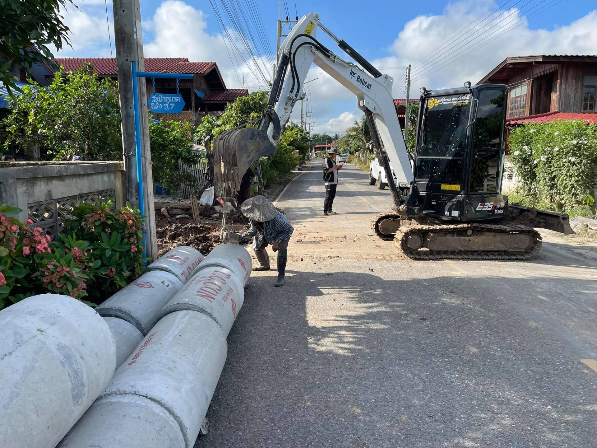 Excavator and worker repairing a road beside stacked concrete pipes on a sunny residential street