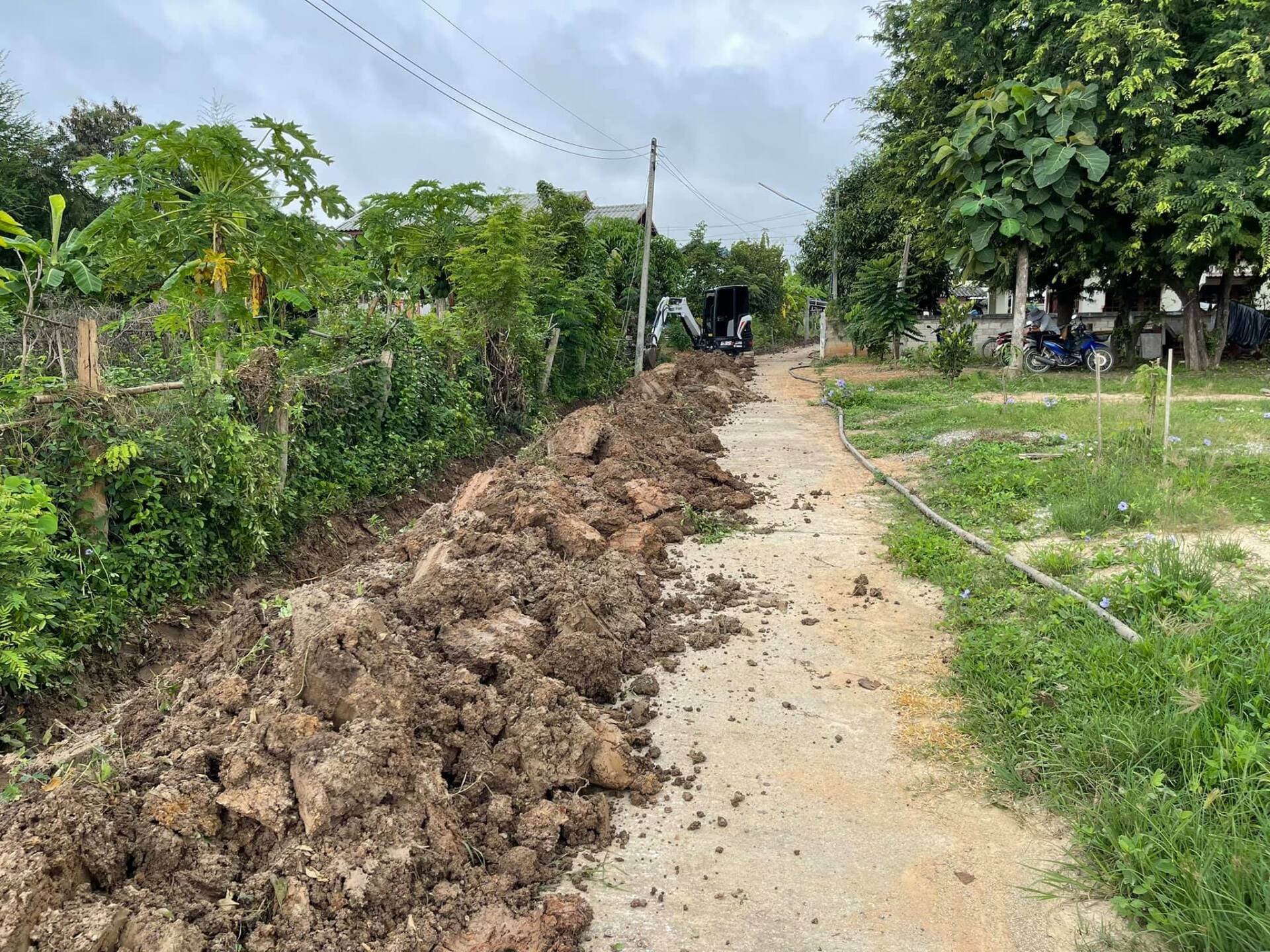 Mud-covered path beside a grassy roadside under cloudy skies; excavated soil piled along the left side.