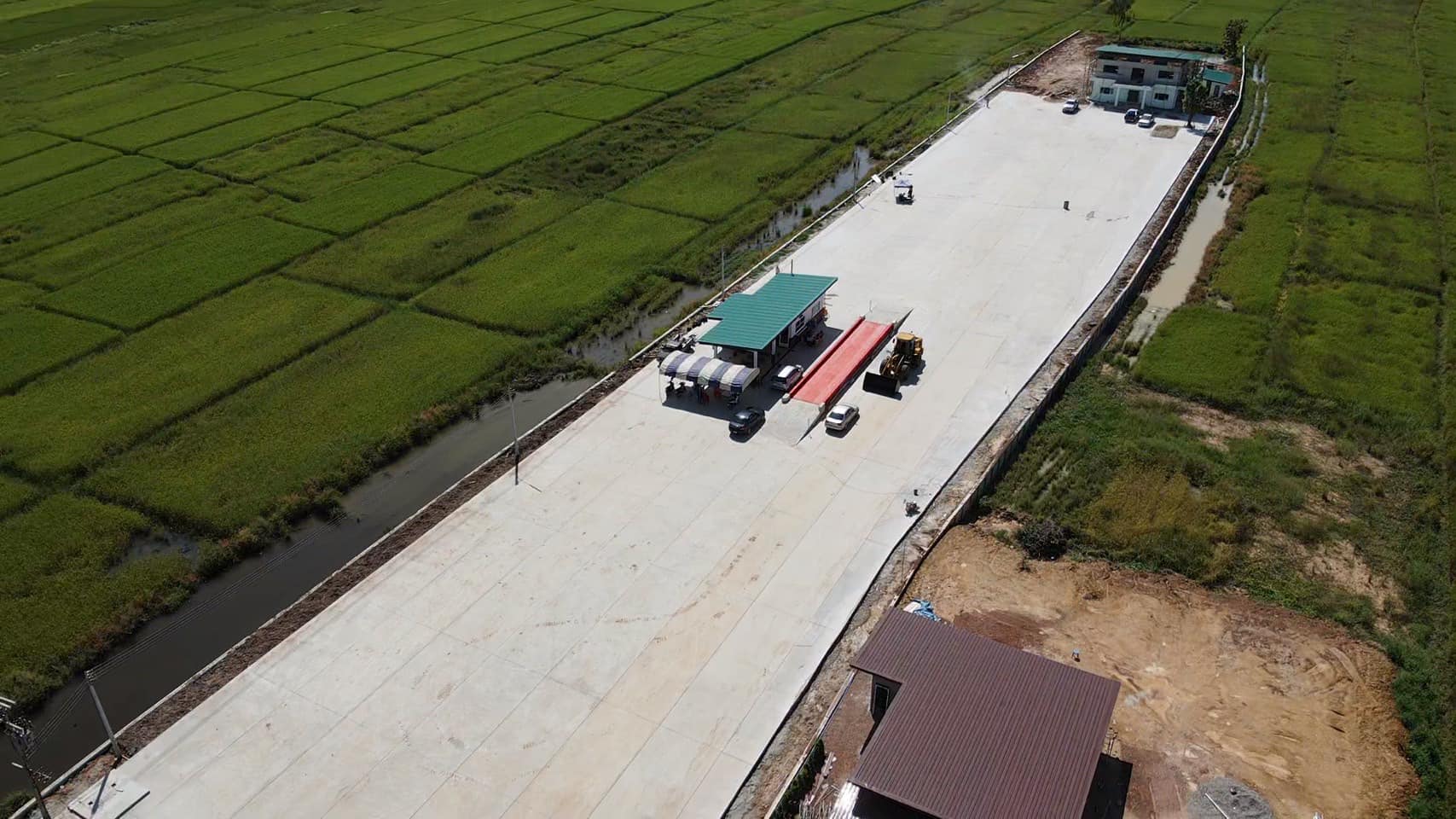 Aerial view of a long concrete road under construction beside green fields, with a truck and workers on site