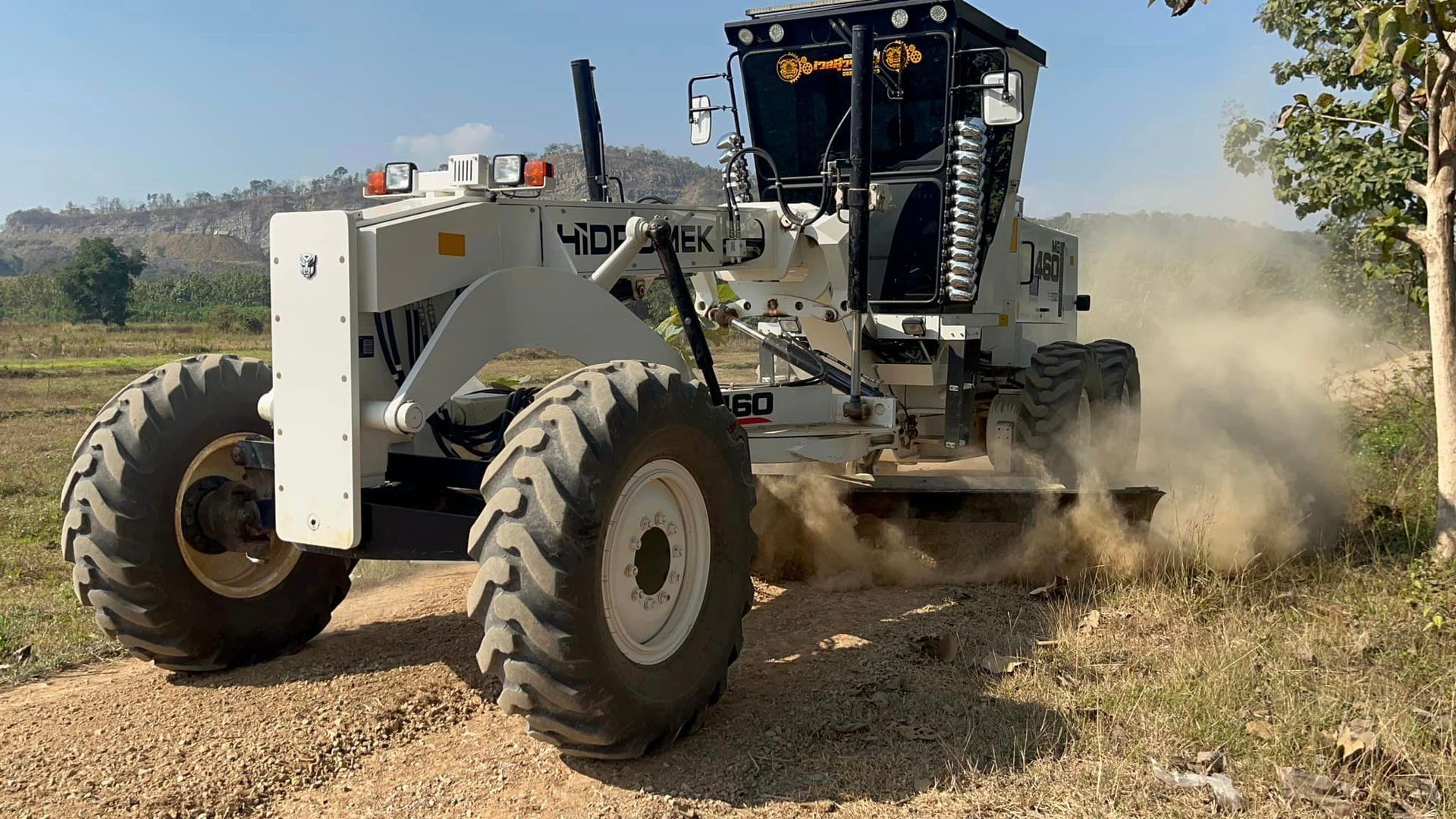 White and black tractor grading a dirt road, kicking up dust in a dry field.