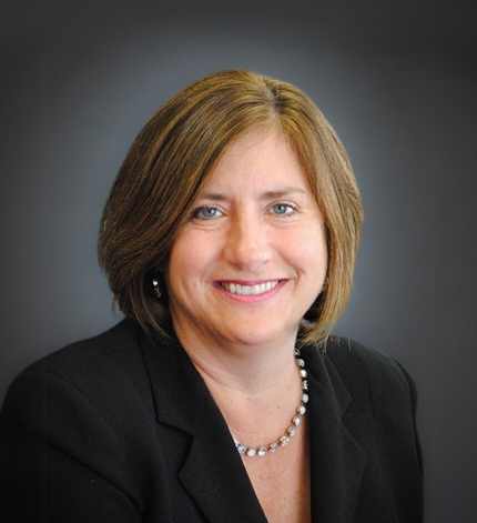 Woman with brown hair smiles, wearing a black blazer and pearl necklace, against a dark background.