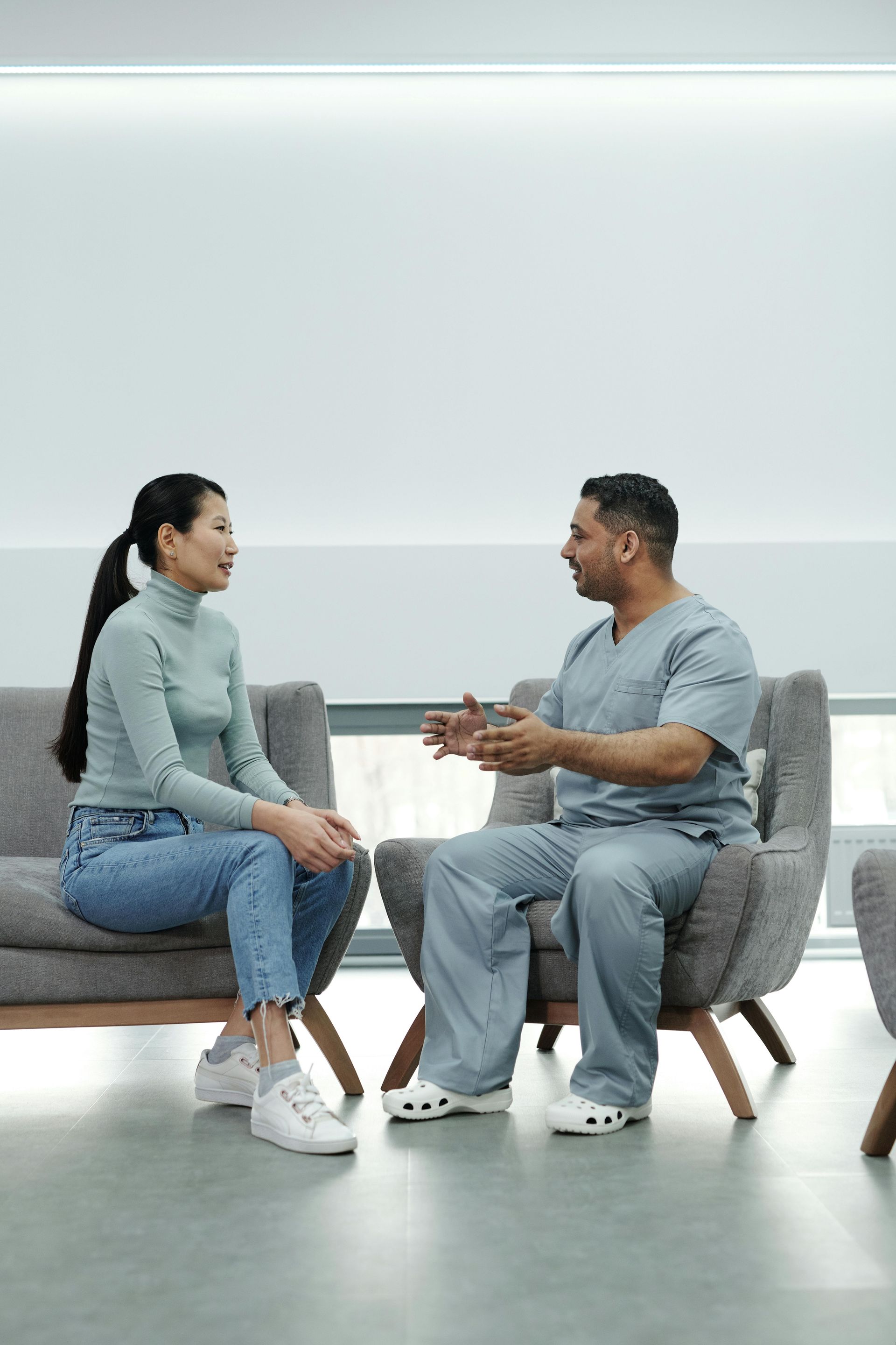 Woman in casual clothes and doctor in scrubs sit facing each other in armchairs, having a conversation.