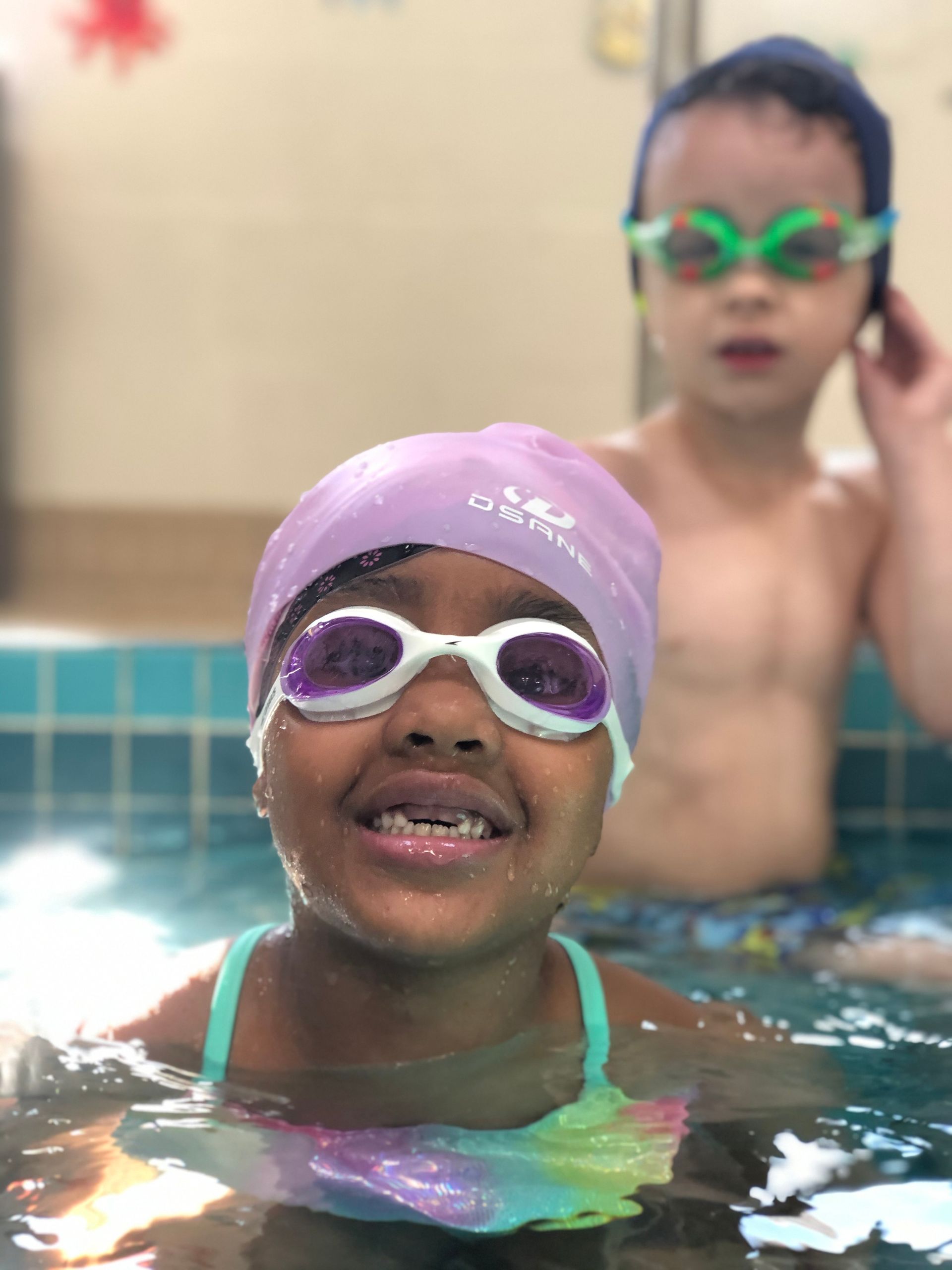 Child in a pool wearing a purple swim cap and goggles, smiling near another child in the background.