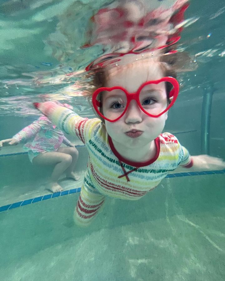 Child underwater in a pool wearing red heart-shaped goggles and a striped swimsuit, reaching forward.