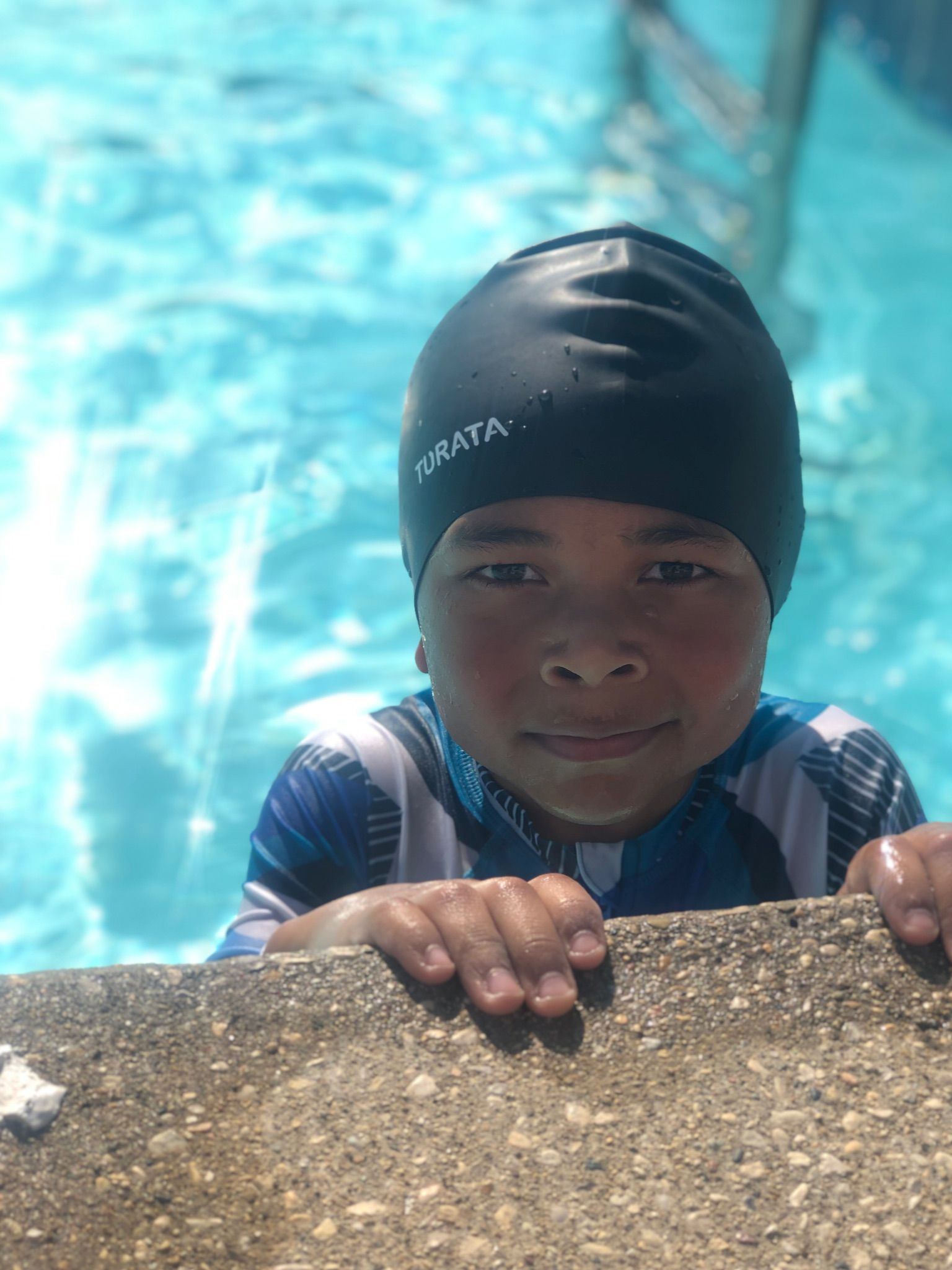 Child in a black swim cap peeks over a pool edge, smiling in bright blue water.