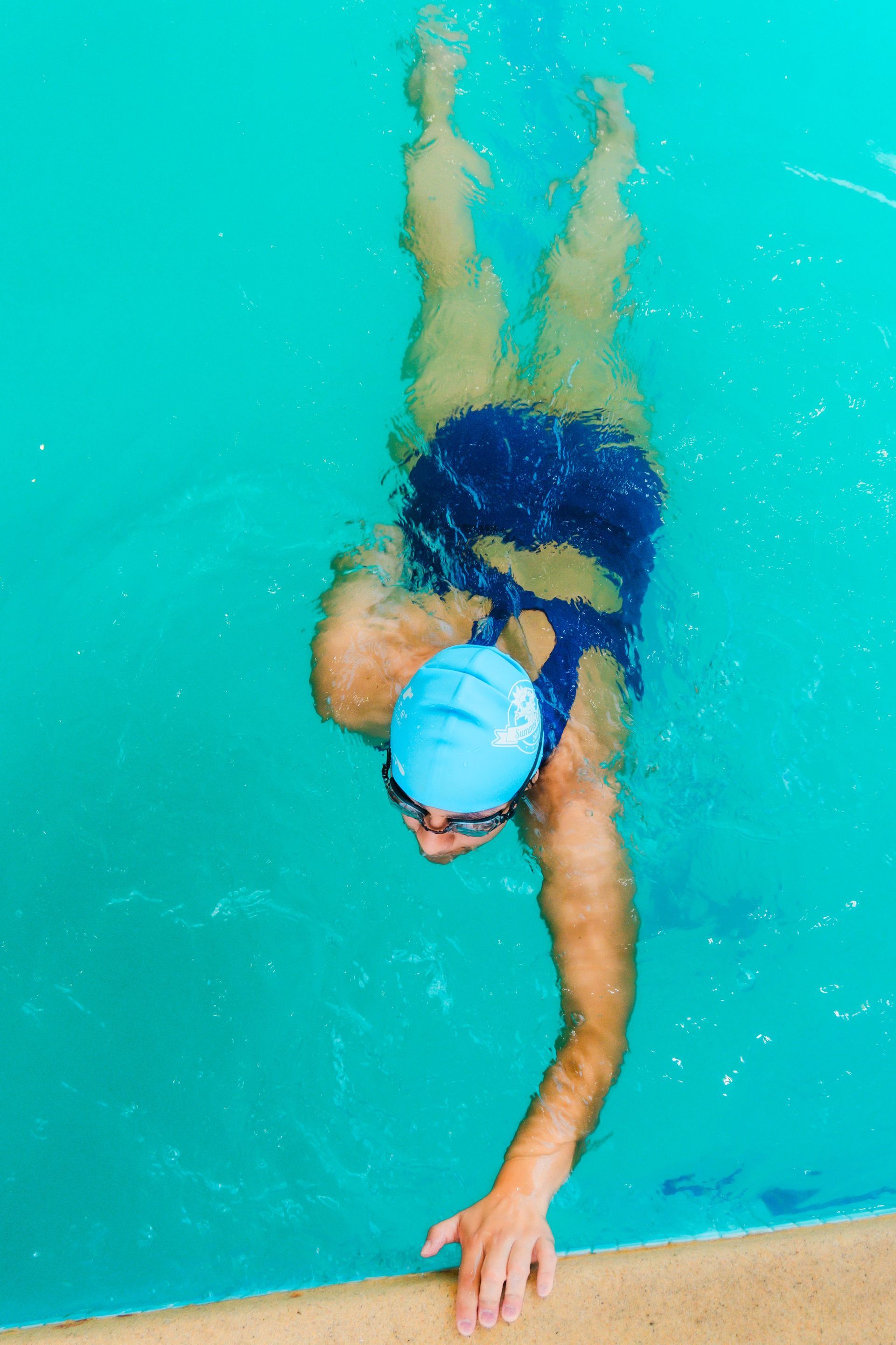 Swimmer reaching a hand along the pool edge in turquoise water, wearing a swim cap and goggles
