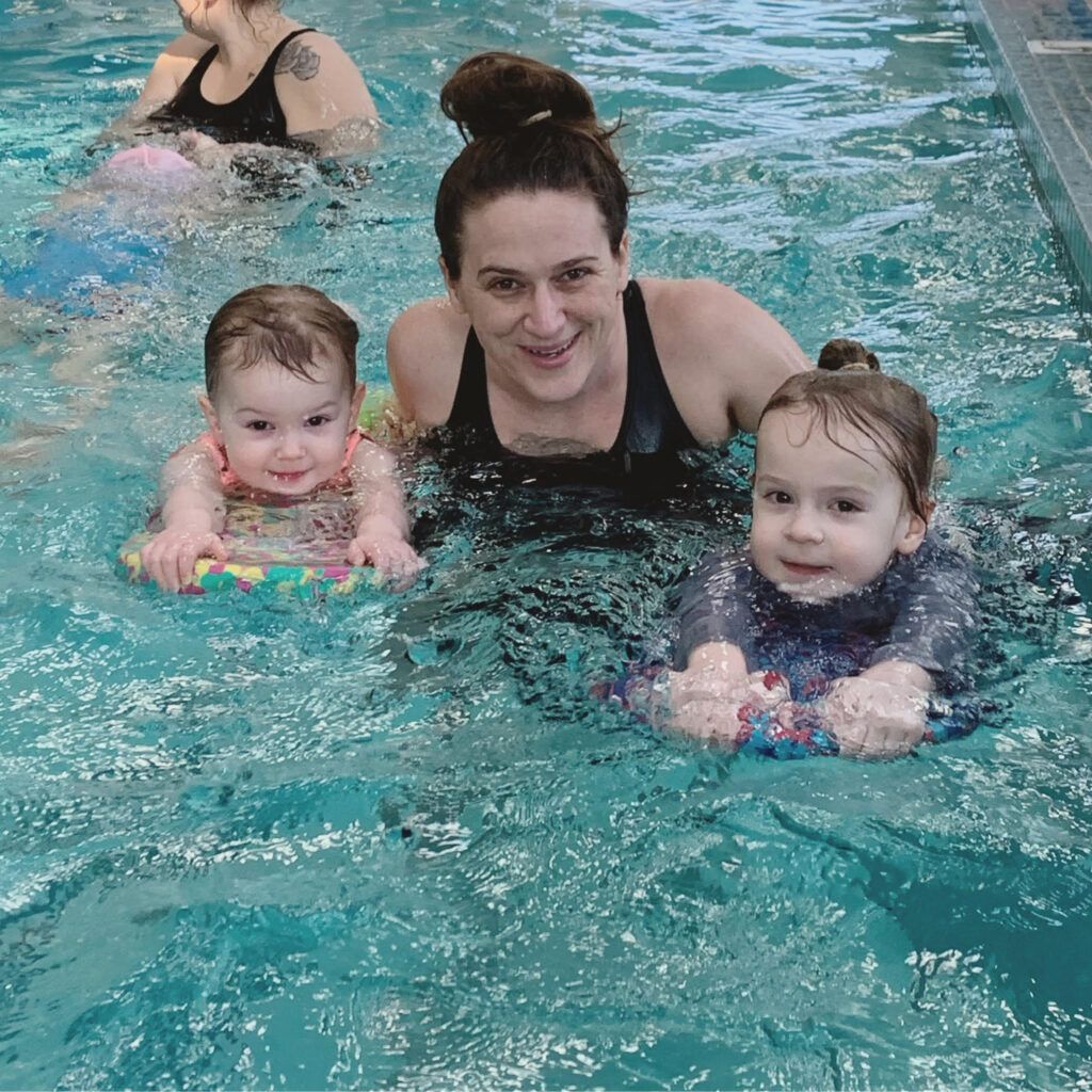 Three swimmers in a pool, smiling and holding onto kickboards, with another swimmer in the background
