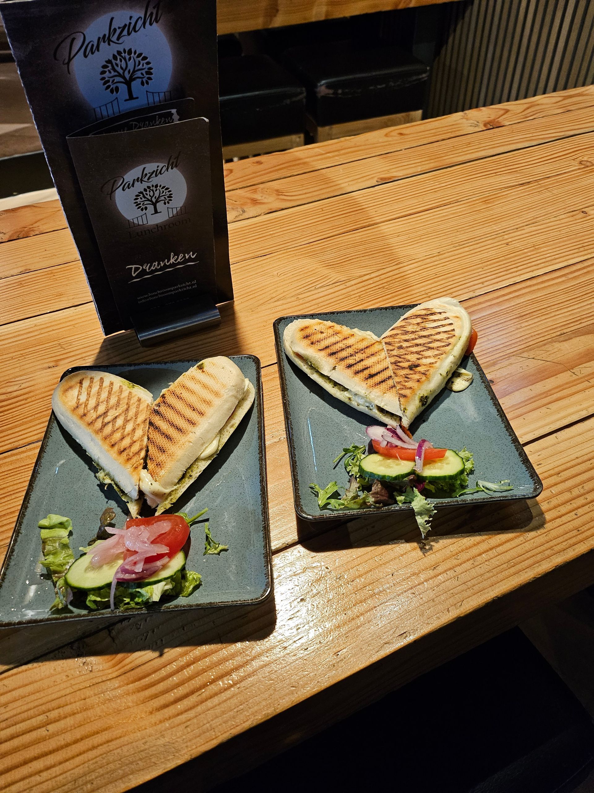 Twee hartvormige panini's op grijze borden, met groenten en een menukaart van het restaurant op een houten tafel.