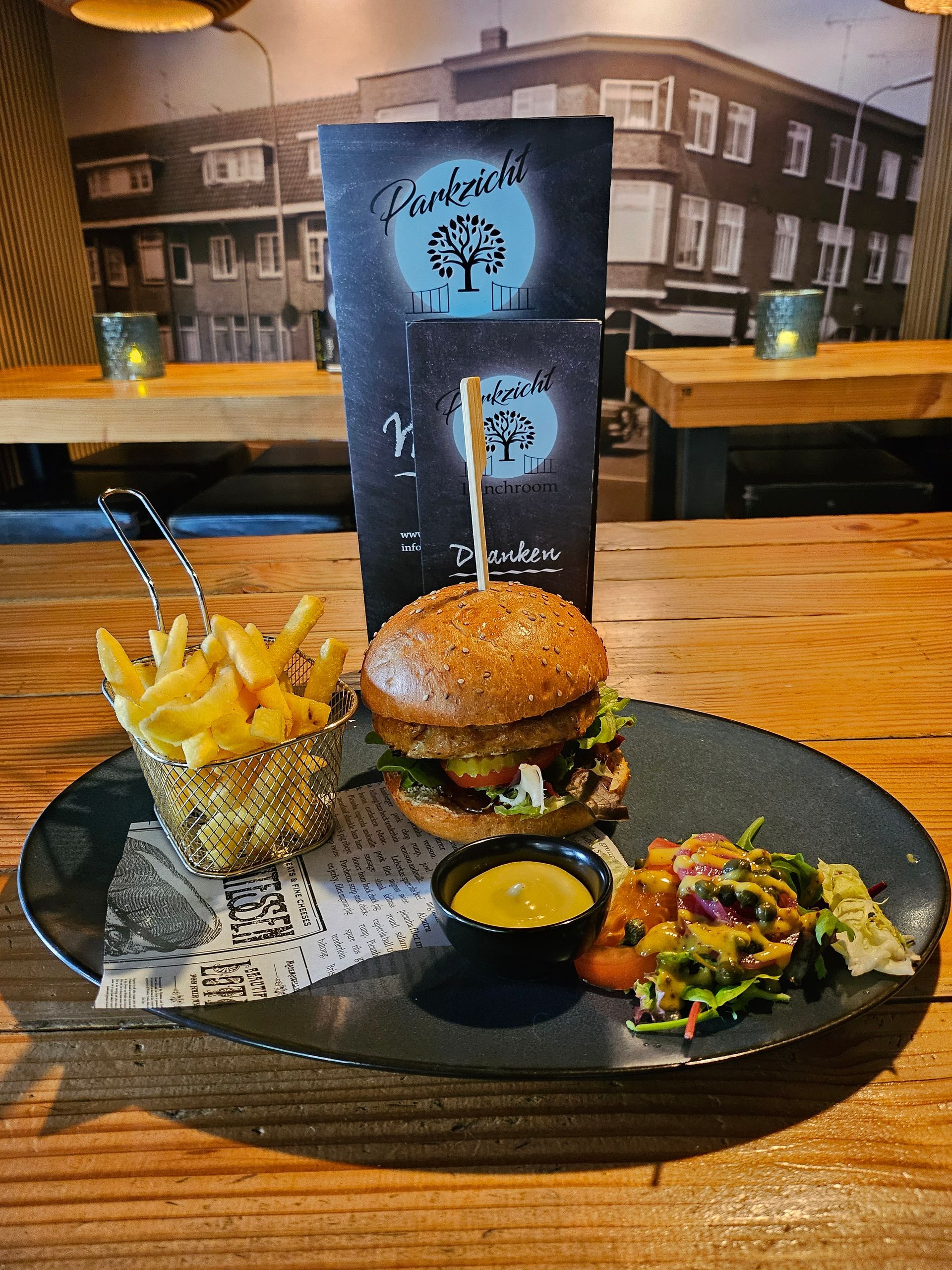 Een hamburger met friet en salade op een bord. Restaurantsetting met menukaart en een gebouw op de achtergrond.