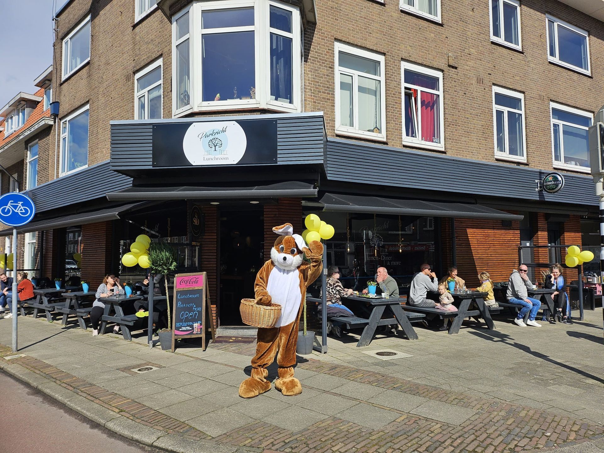 Een persoon in een konijnenpak met ballonnen staat voor een restaurant. Mensen zitten aan tafels buiten.