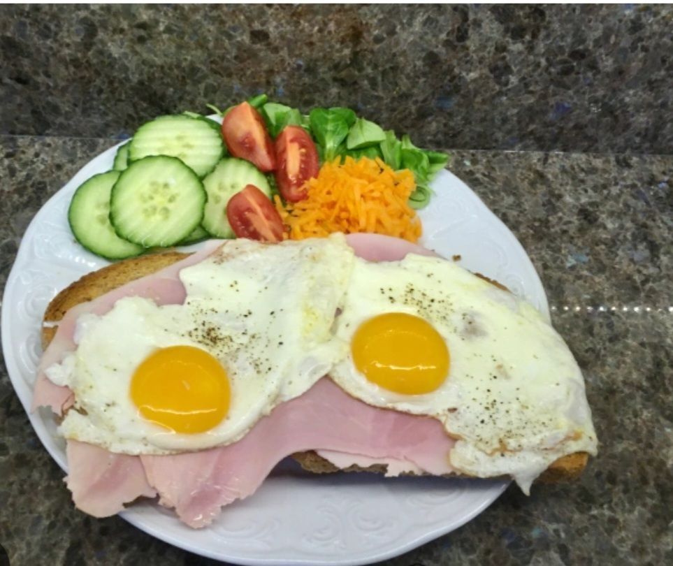 Toast met ham en ei, geserveerd met een salade van komkommer, tomaat, wortel en sla op een wit bord.