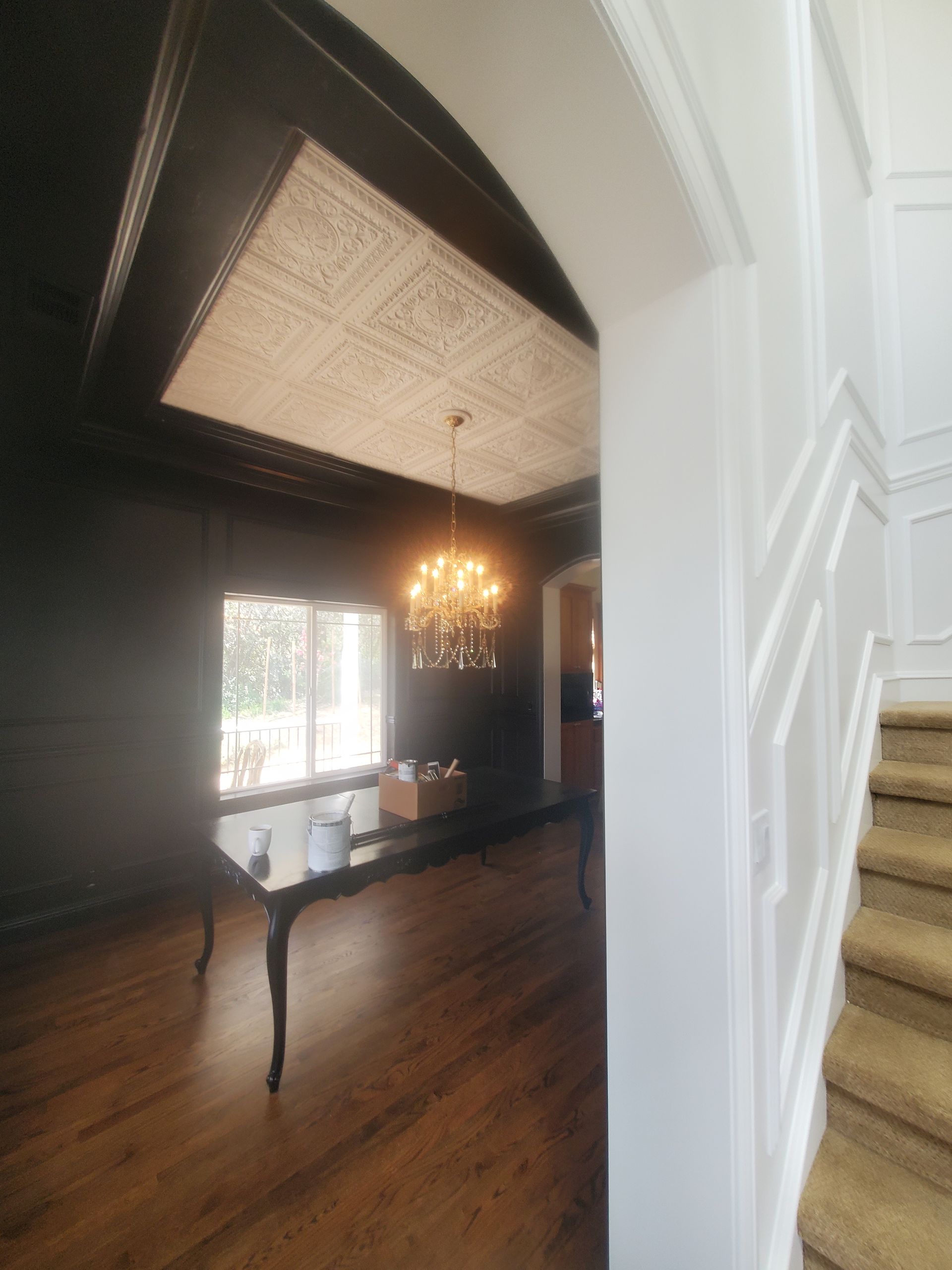 Interior shot of a dark-painted dining room with ornate ceiling design, chandelier, and a view into a hallway with a staircase.
