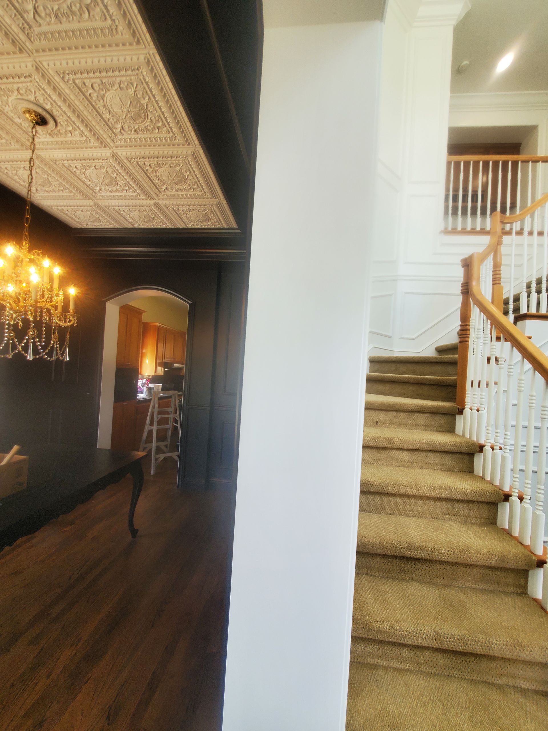 Interior view: Stairs with brown carpet, white banister, and a chandelier in a dining room with dark walls and ceiling.