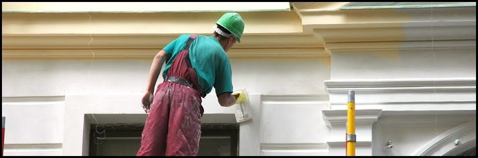 Painter in a green hard hat and red overalls rolling white paint onto an exterior wall.