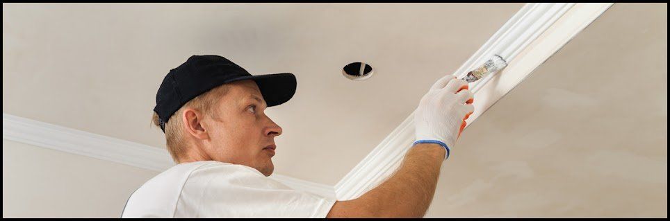 A person in a black cap paints trim near a ceiling light.