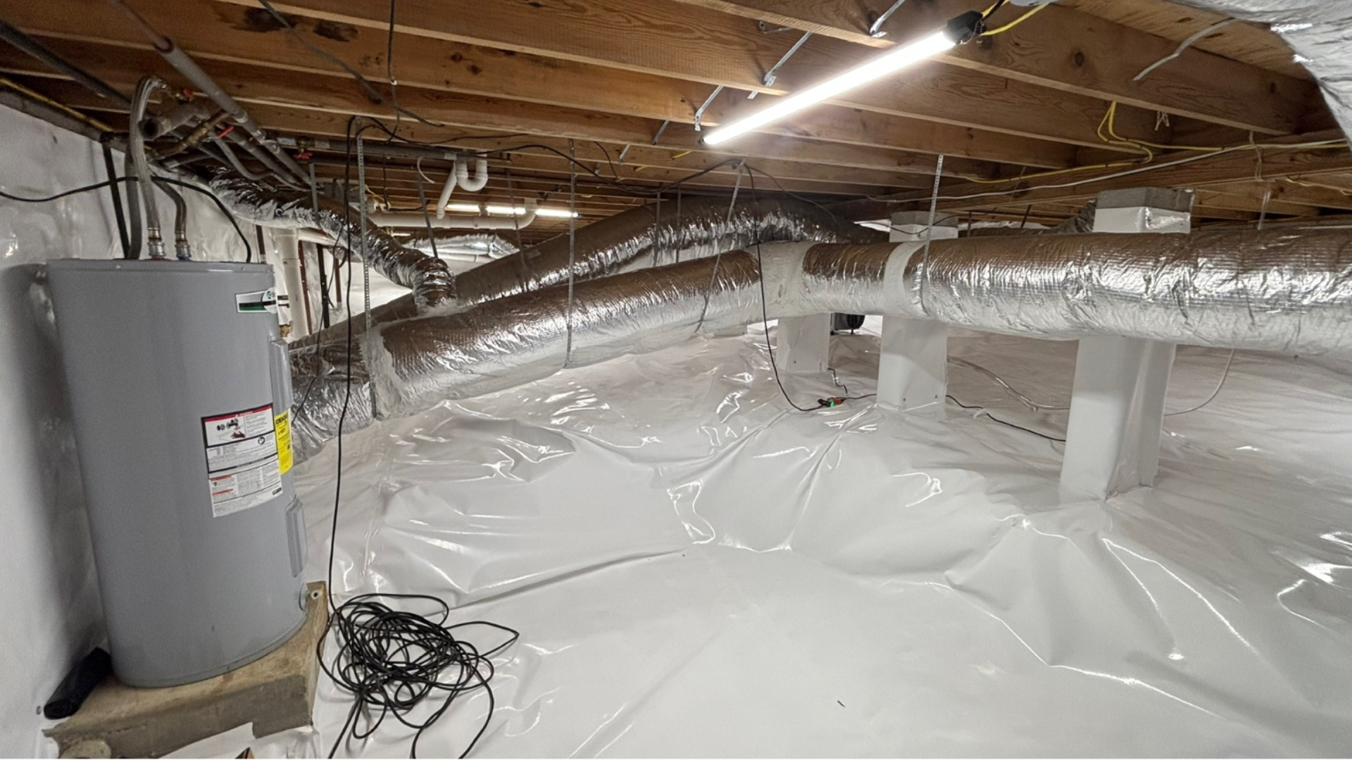 Crawlspace with white vapor barrier flooring, exposed beams, ductwork, and a water heater in the corner