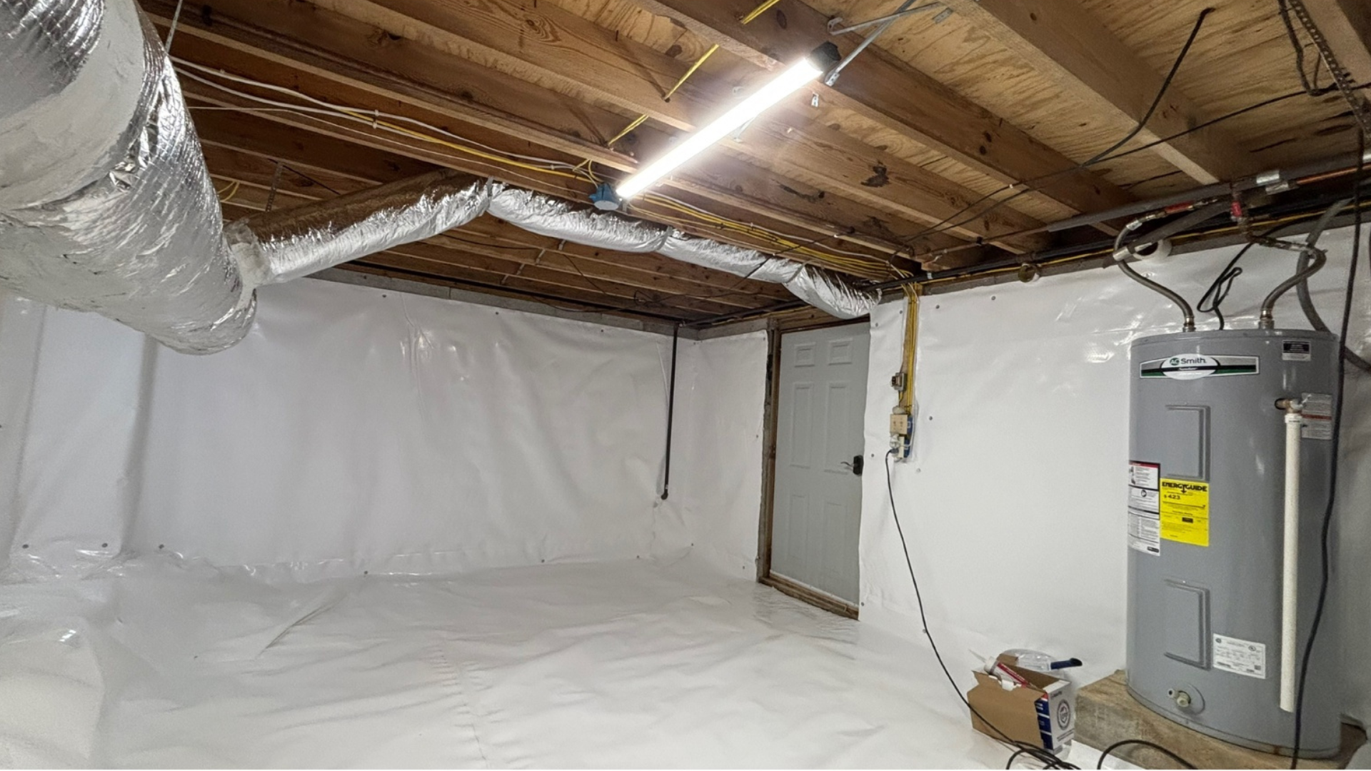 Plastic-lined basement with exposed beams, ductwork, and a water heater or furnace on the right.