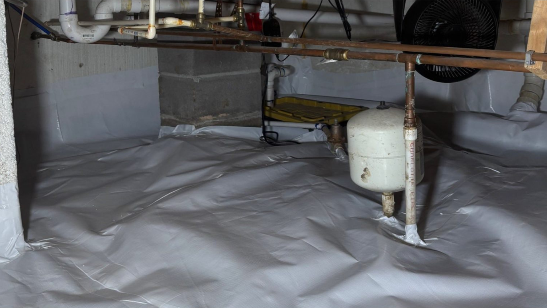 Plastic-covered crawlspace with pipes and a white tank under exposed joists