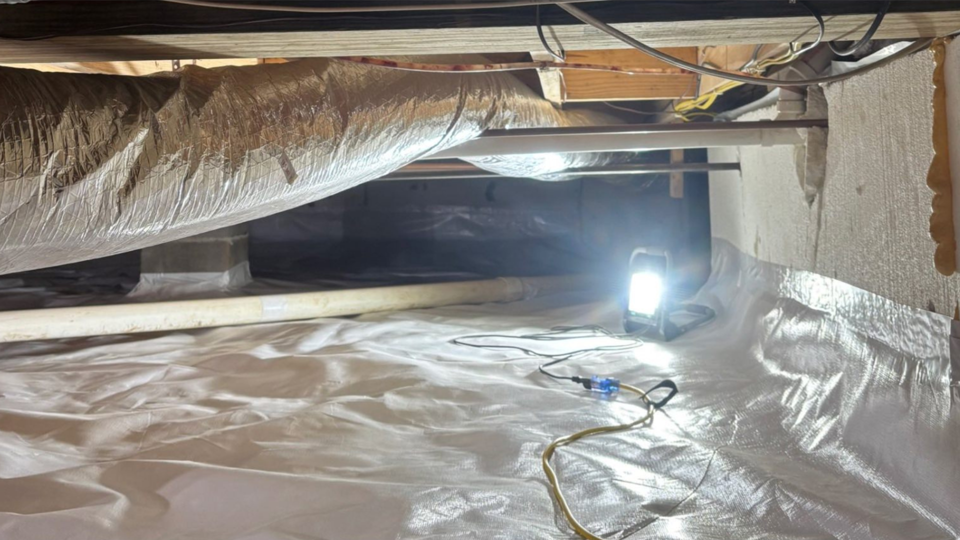 Insulated attic crawlspace with exposed beams, plastic sheeting, and a bright work light on the floor