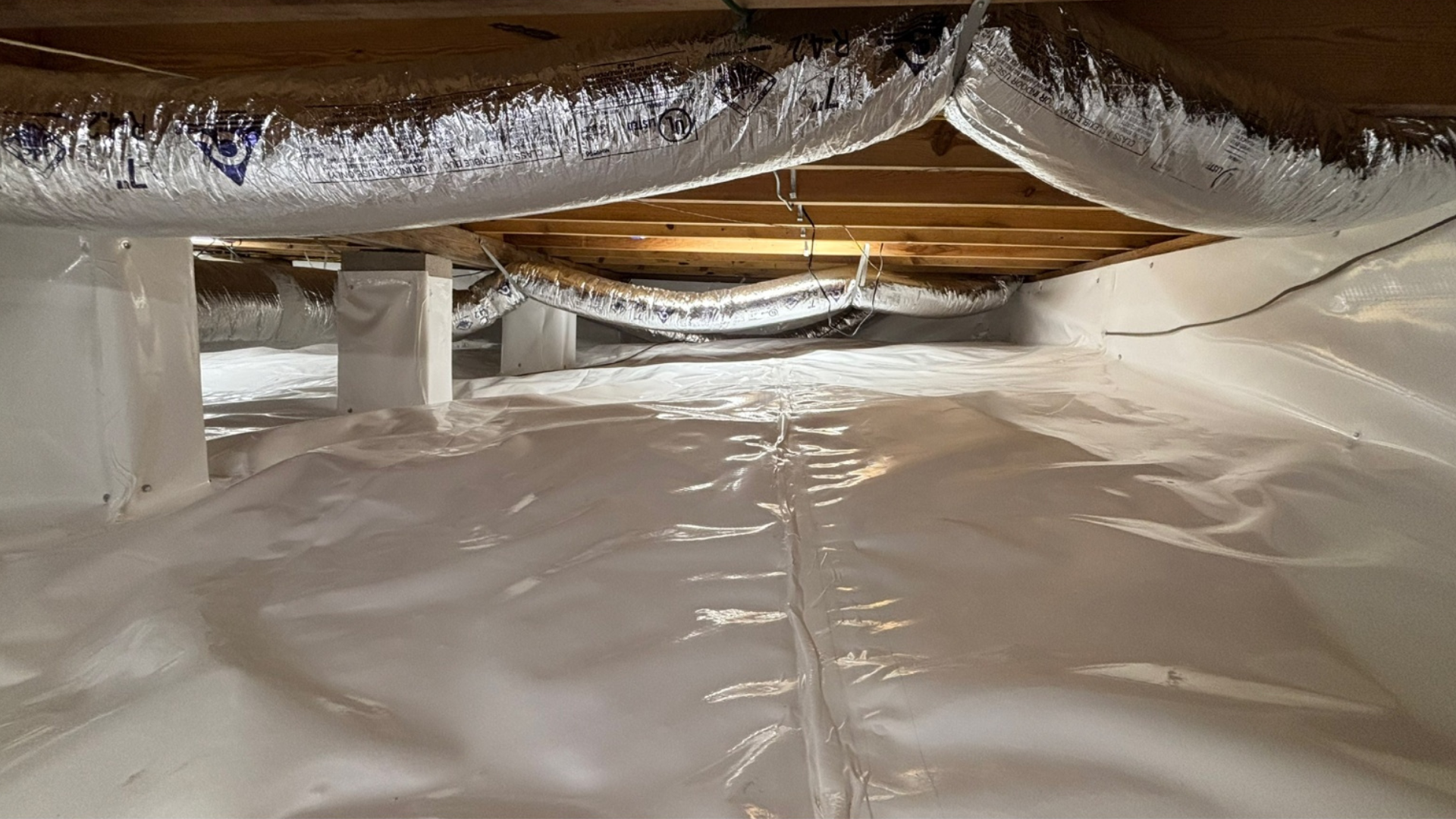 Low attic crawlspace lined with white vapor barrier and exposed wooden beams overhead
