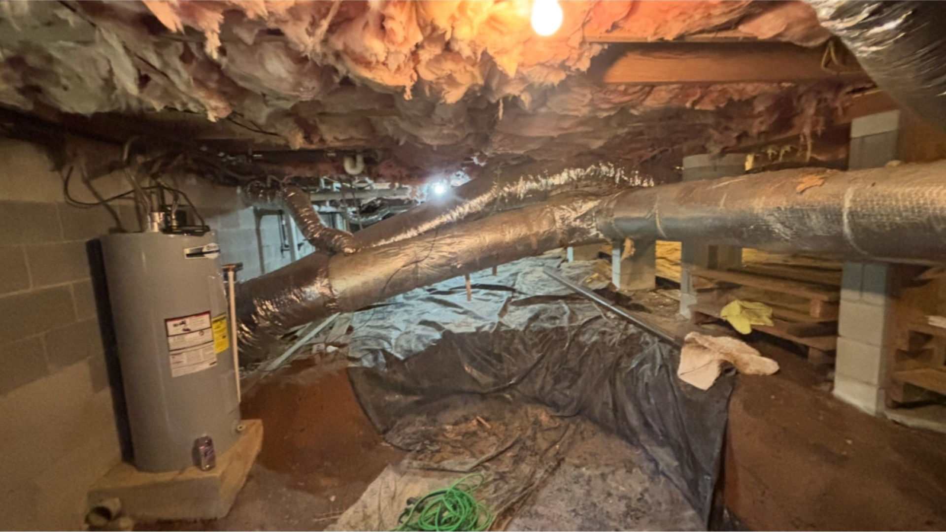 Dim basement crawlspace with exposed pipes, insulation, and a hanging light bulb.