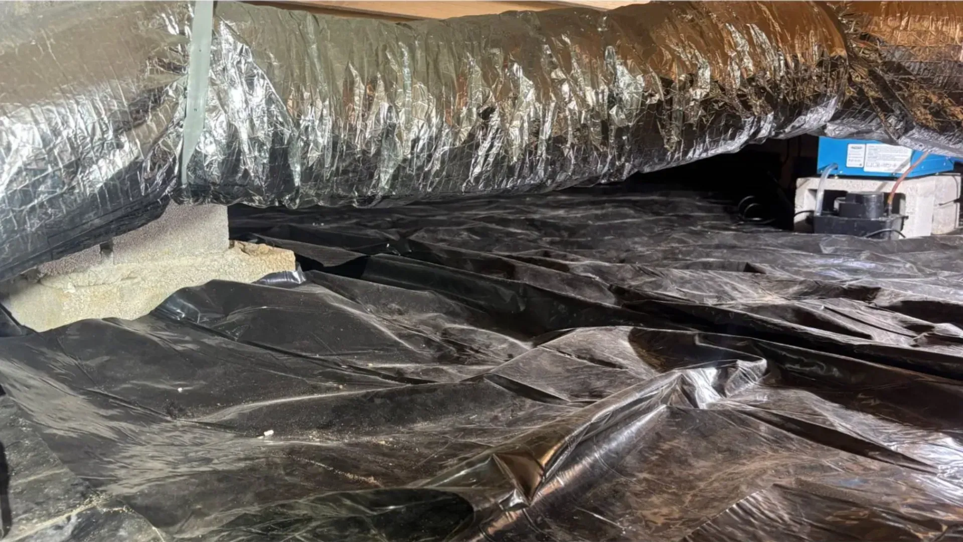 Black plastic sheeting and insulated ducting in a crawl space or attic area, with tools in the background