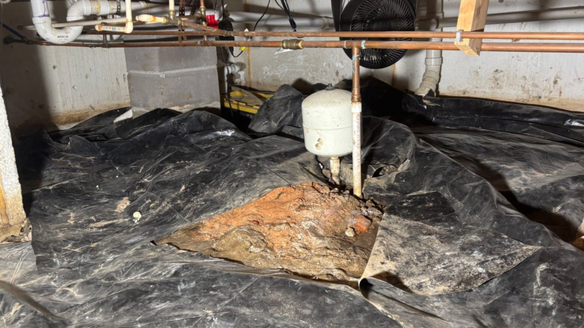 Basement crawlspace with black plastic vapor barrier, exposed pipes, and a white bucket near a patched floor section